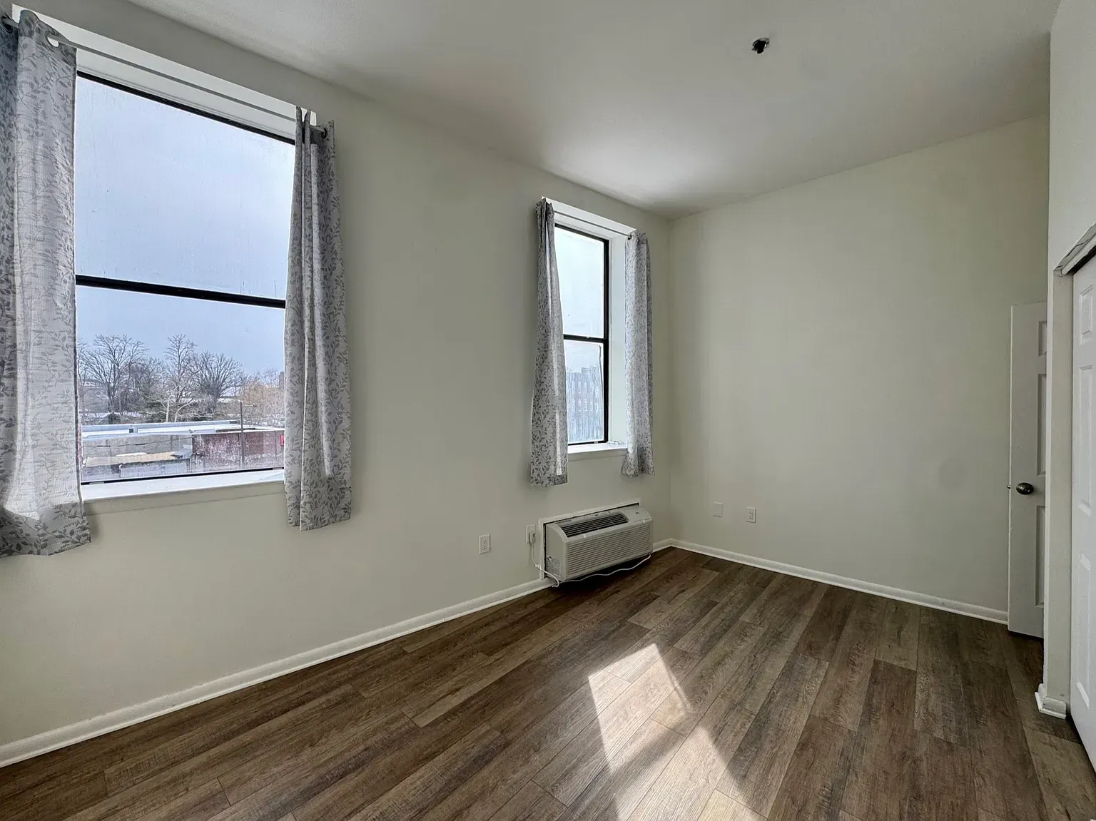 Empty white room with wood floors, two windows, and a wall heater