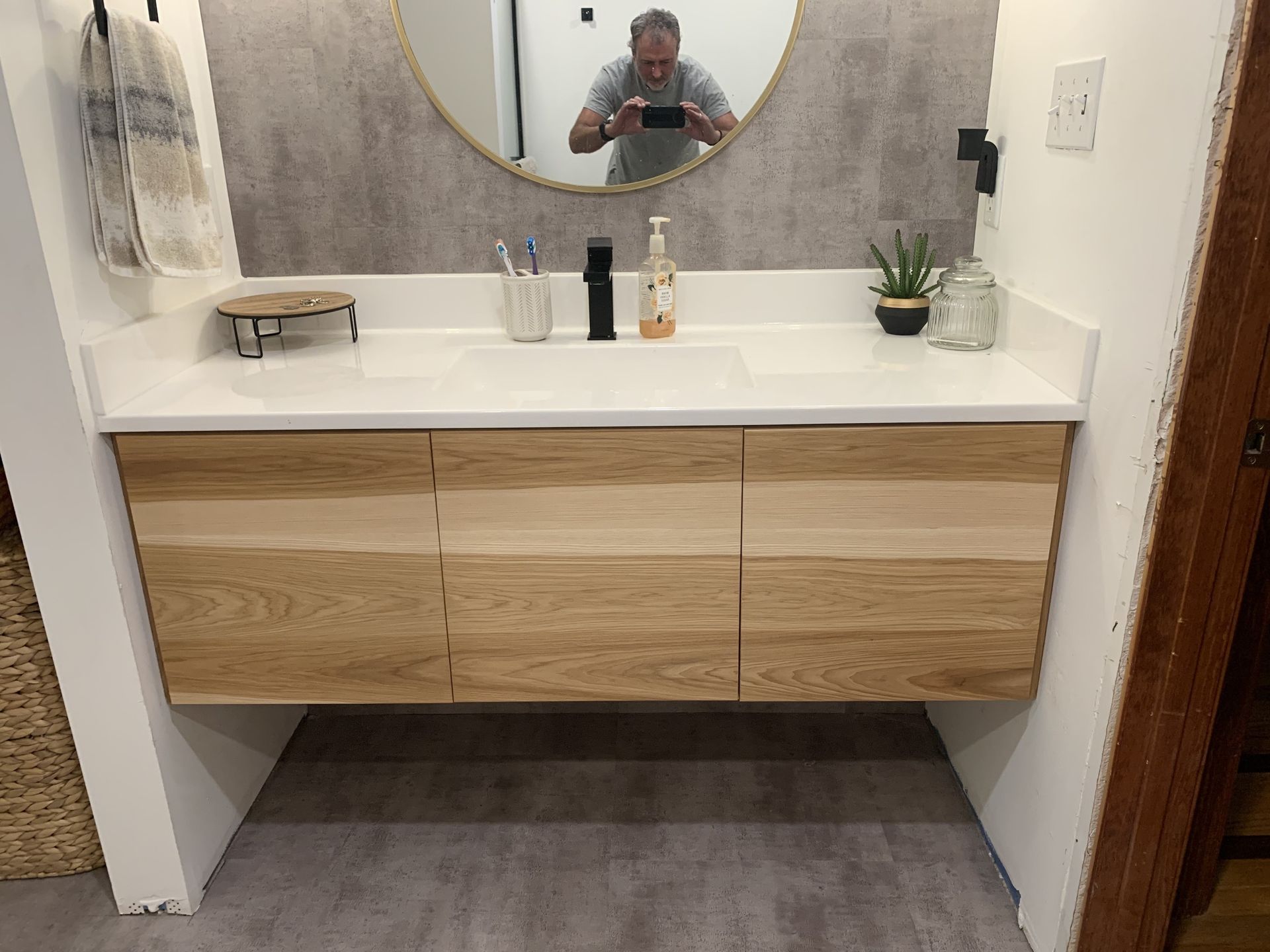 Bathroom with floating light wood vanity, white countertop, round mirror, and person taking a photo.