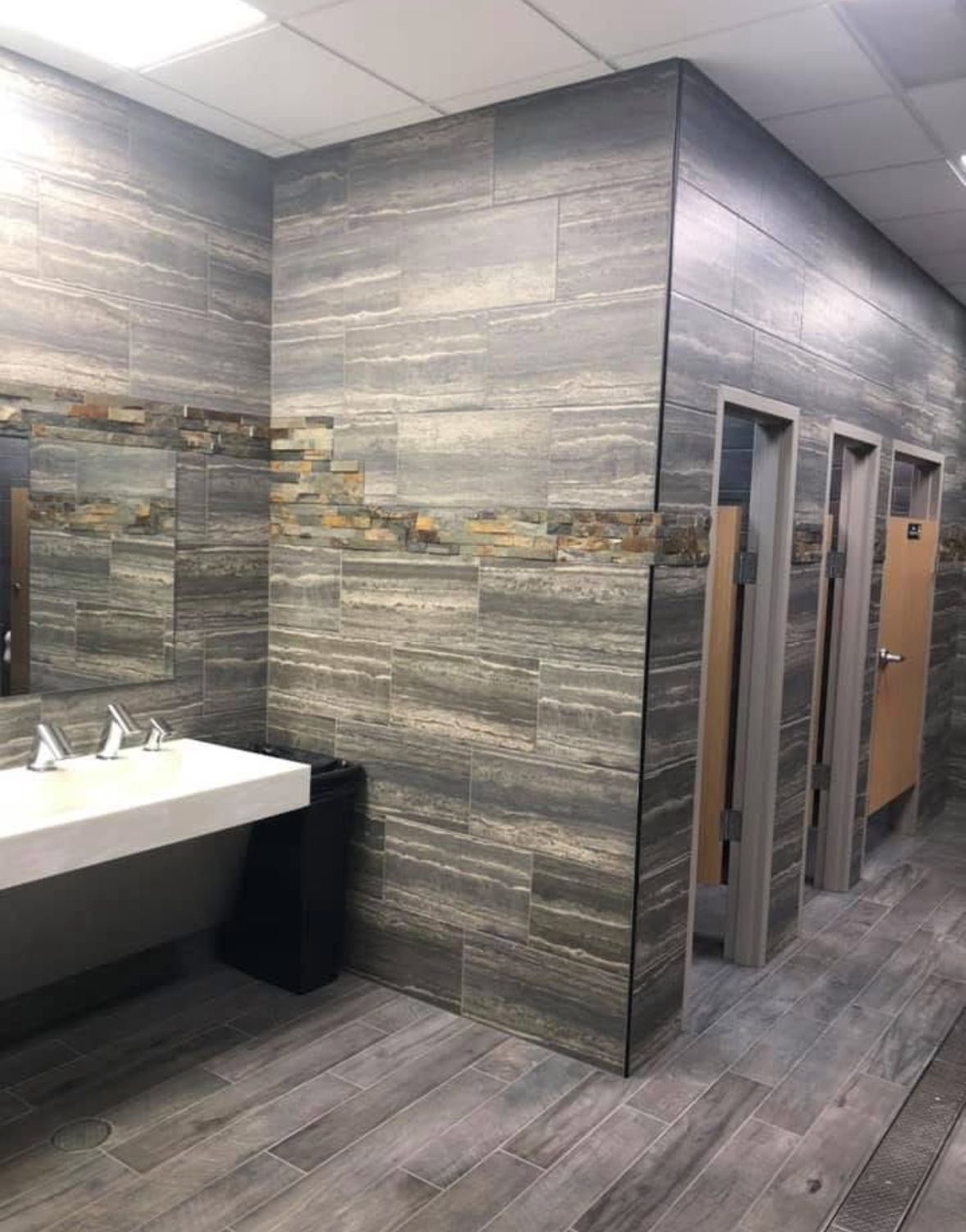 Public restroom with gray and brown tile, three stalls, and a white sink.
