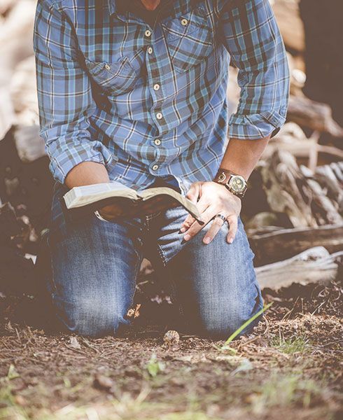 A man is kneeling down in the dirt reading a bible.
