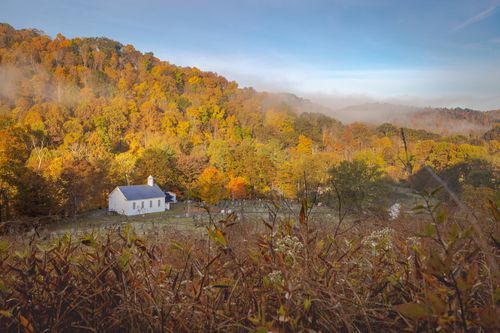 A white church building is sitting on top of a hill in the middle of an autumn forest.