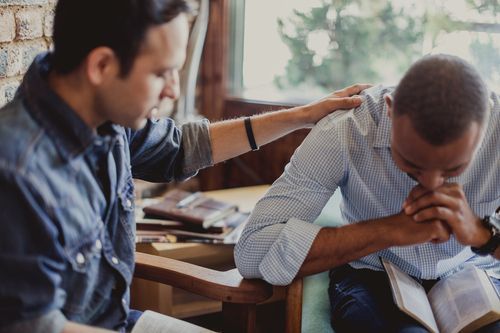A man is putting his hand on another man 's shoulder while they pray together.
