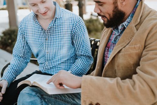Two men are sitting on a bench reading a Bible.