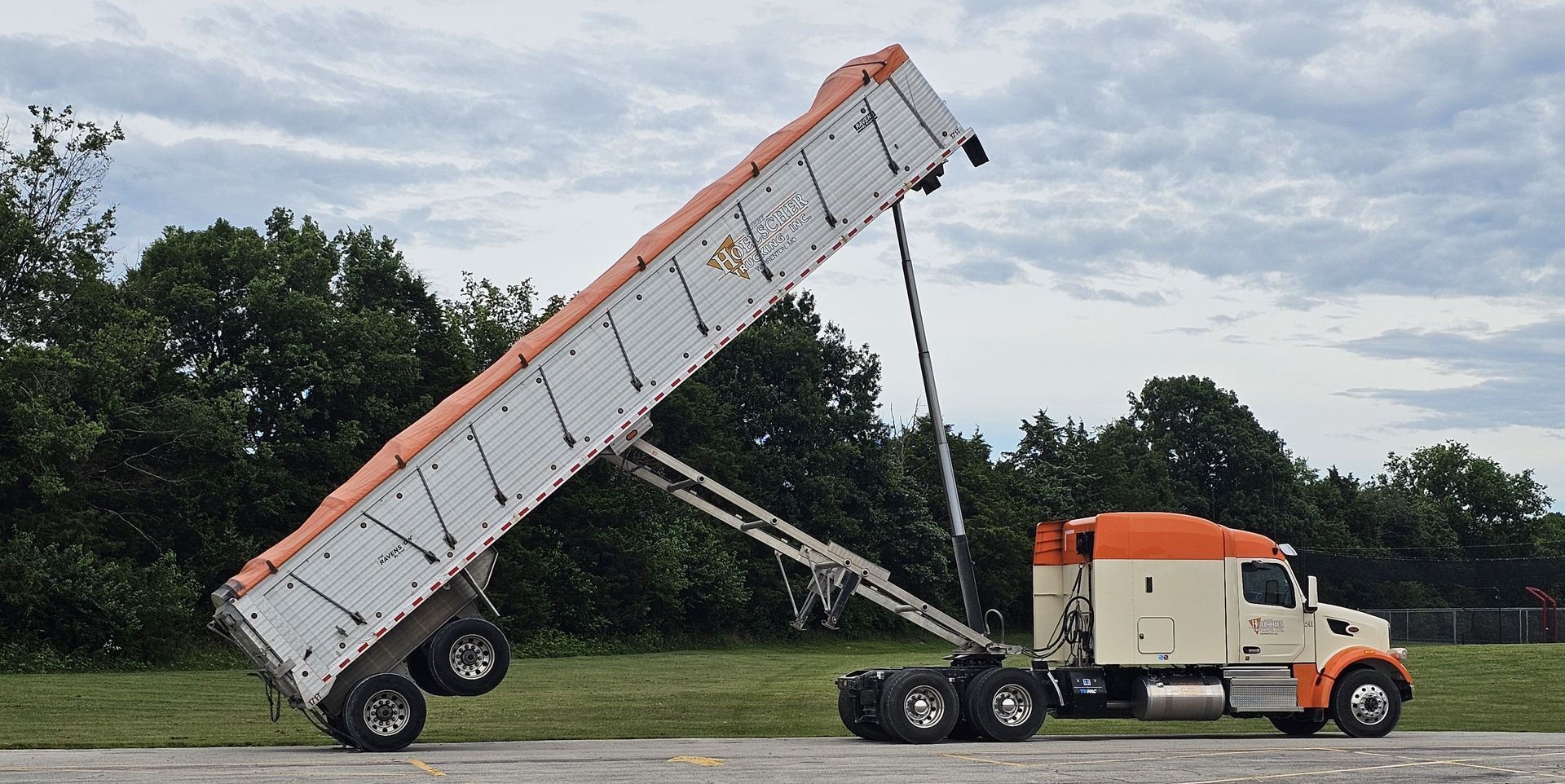 Semi-truck with a lifted cargo trailer, preparing to unload. Beige and orange cab on a grassy area, cloudy sky.