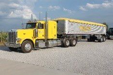 Yellow Peterbilt semi-truck with a silver grain trailer, parked on gravel.