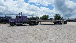 Purple semi-truck with a flatbed trailer parked outdoors on a sunny day with clouds.