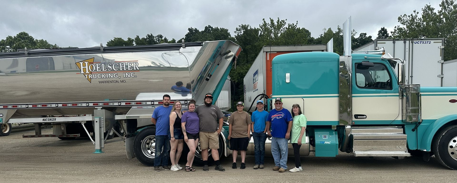 Group of people stand in front of turquoise semi-truck and trailer. Cloudy sky and trees in the background.
