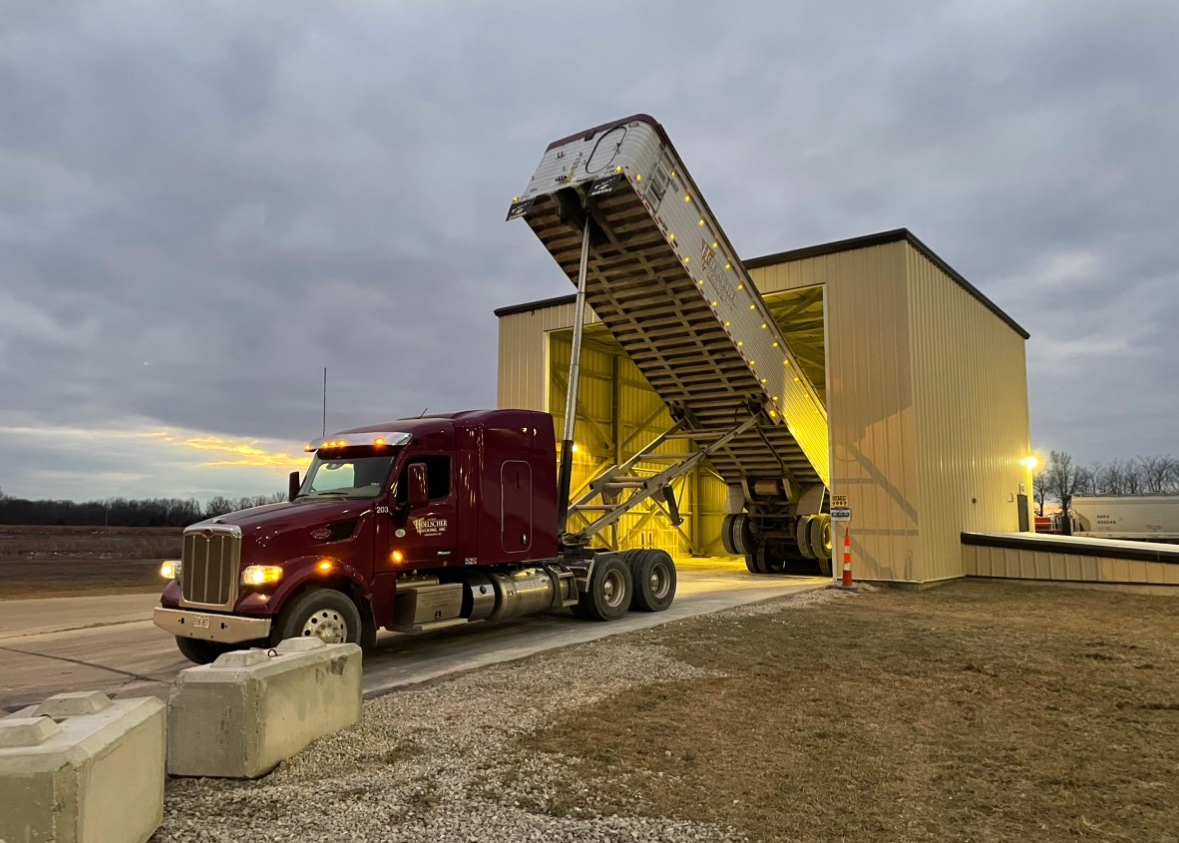 A maroon semi-truck unloading cargo into a raised structure, on a cloudy day.