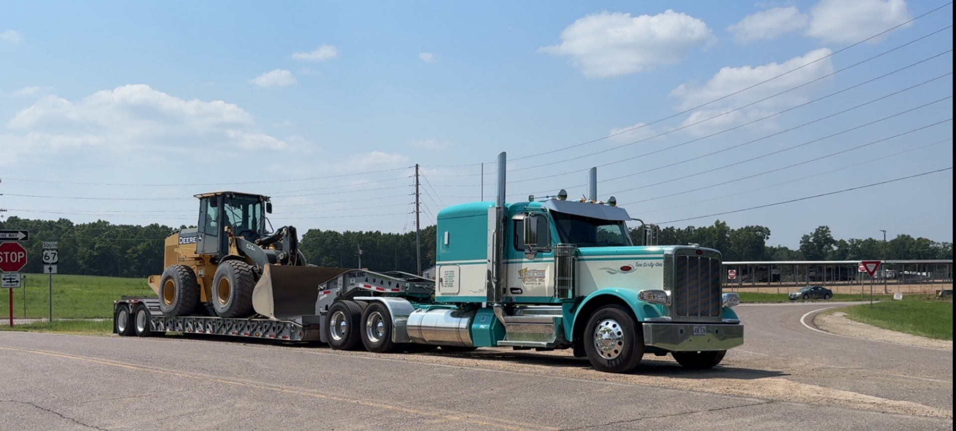 A teal and white semi-truck hauls a front-end loader on a lowboy trailer on a sunny road.
