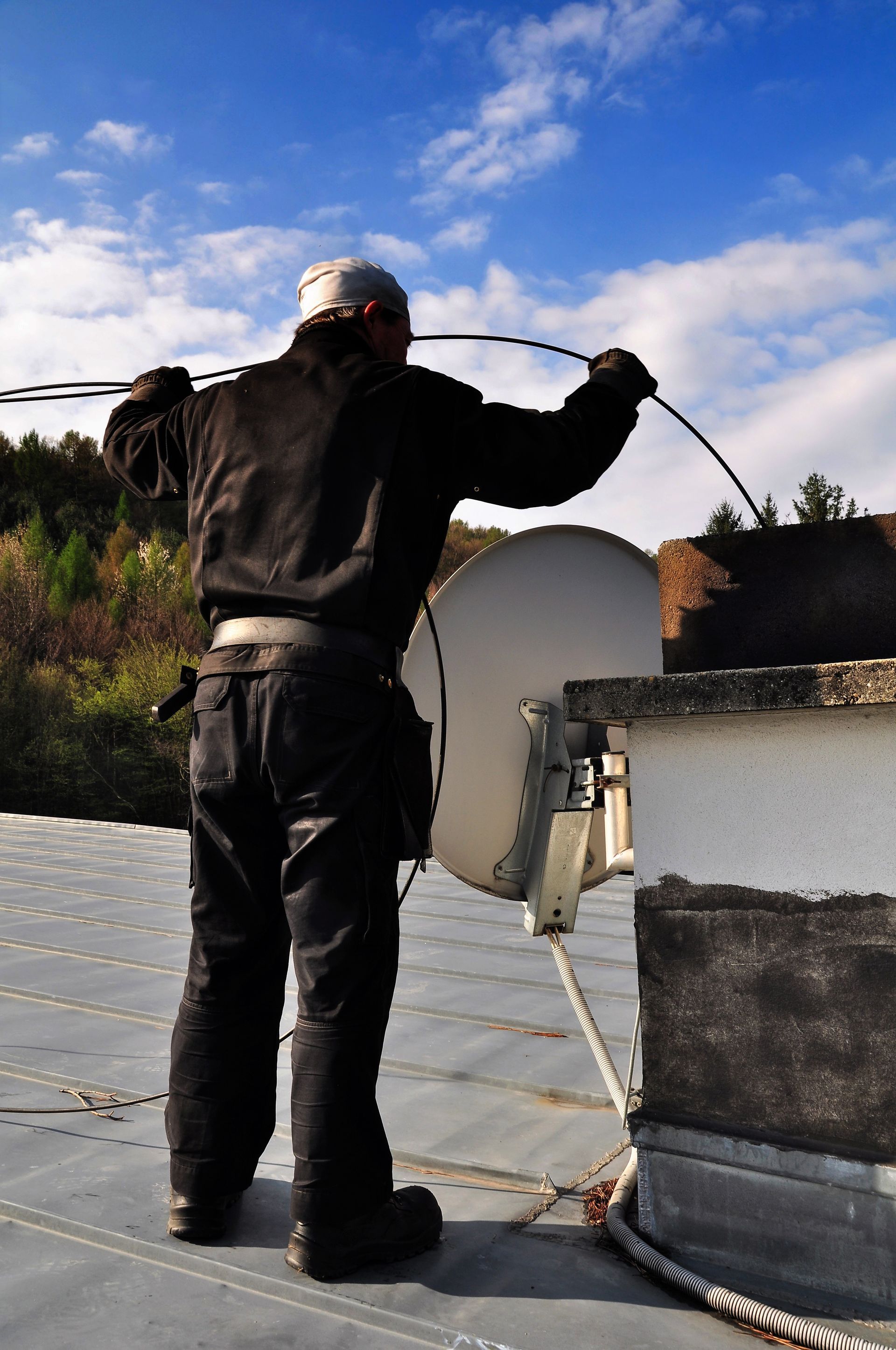 Person on a rooftop working on a satellite dish, holding a cable against a blue sky. Person on a rooftop working on a satellite dish, holding a cable against a blue sky.
