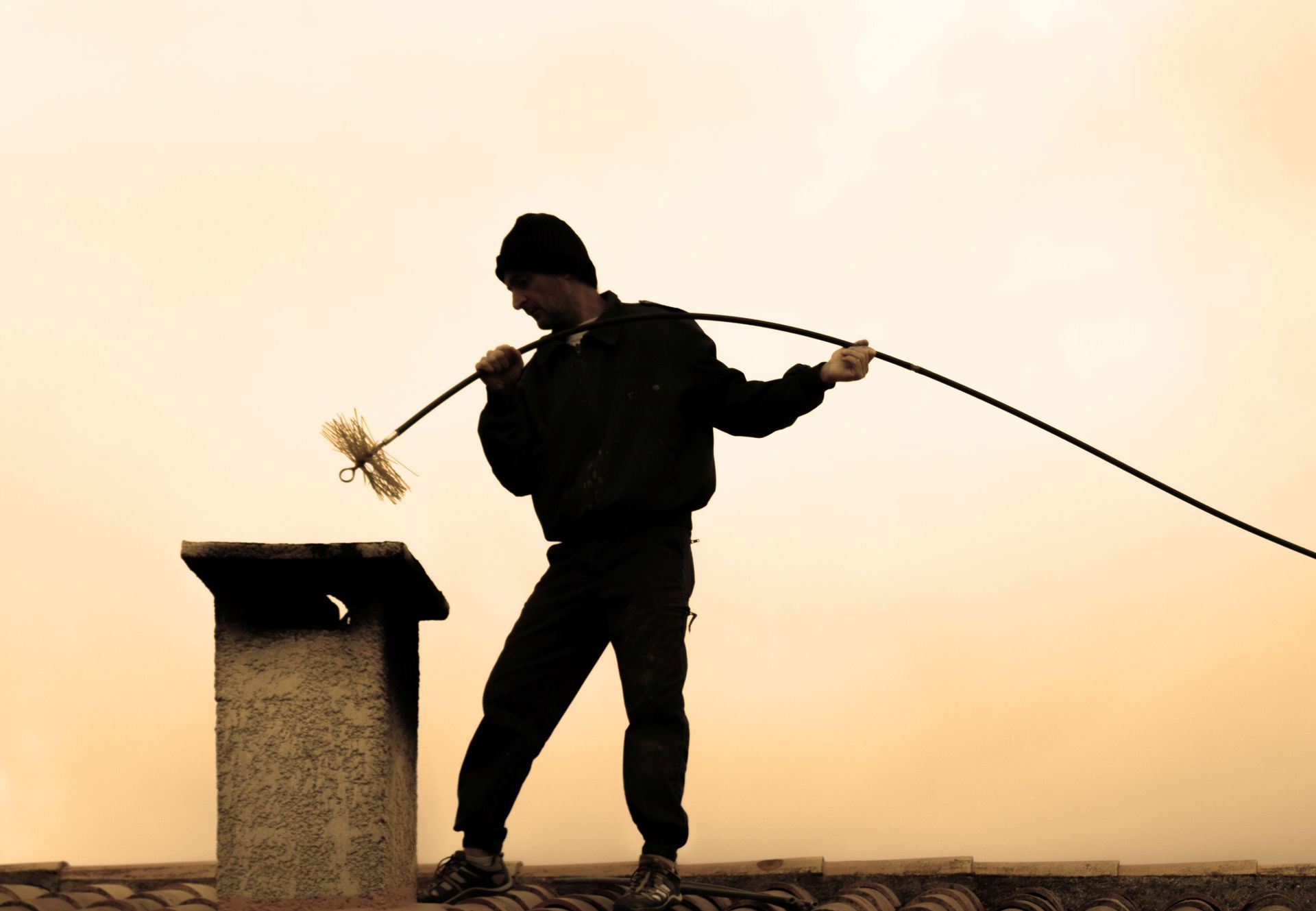 A chimney sweep silhouette on a rooftop cleaning a chimney at sunset.
