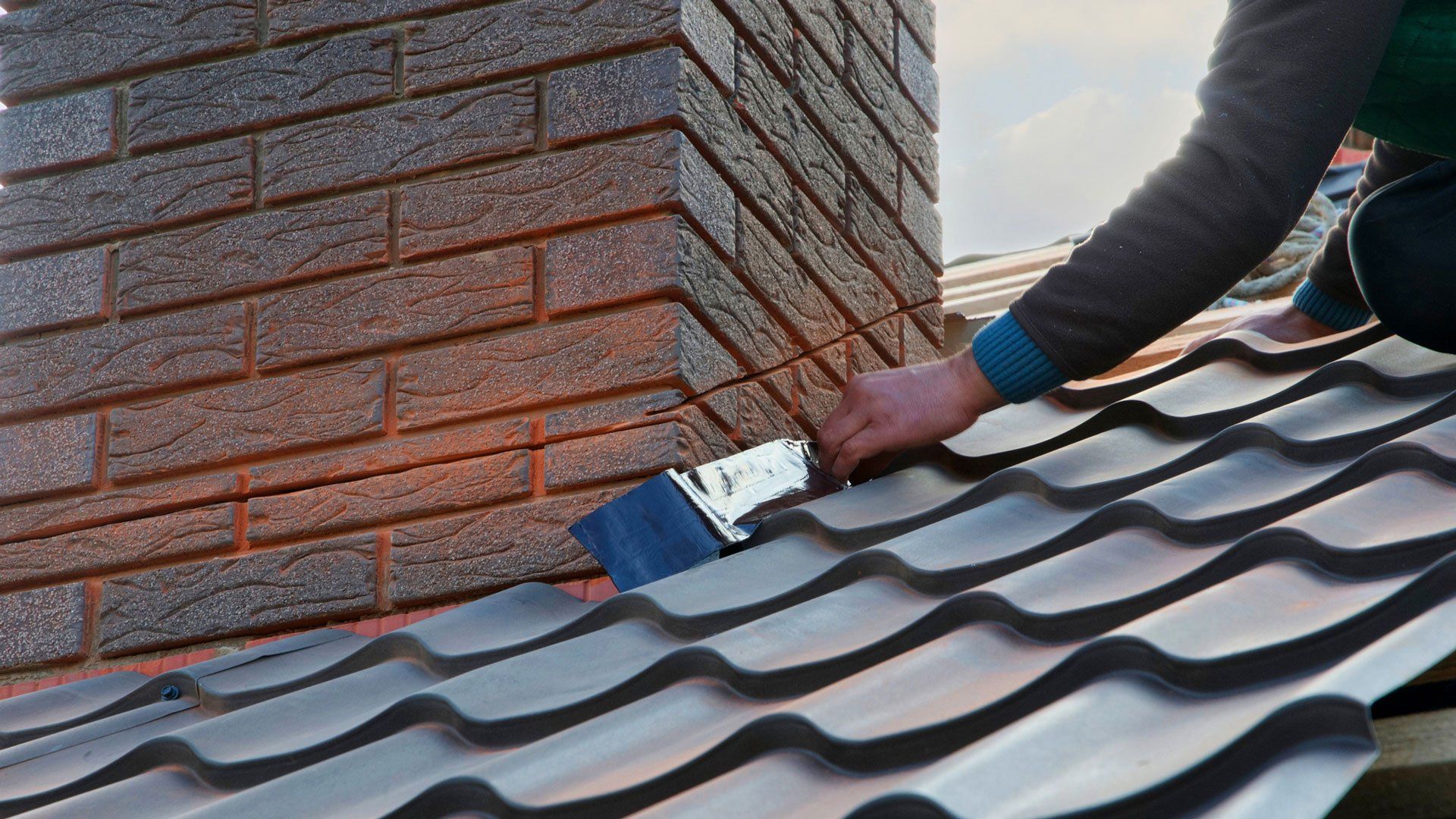 Person installing flashing around a brick chimney on a metal roof.
