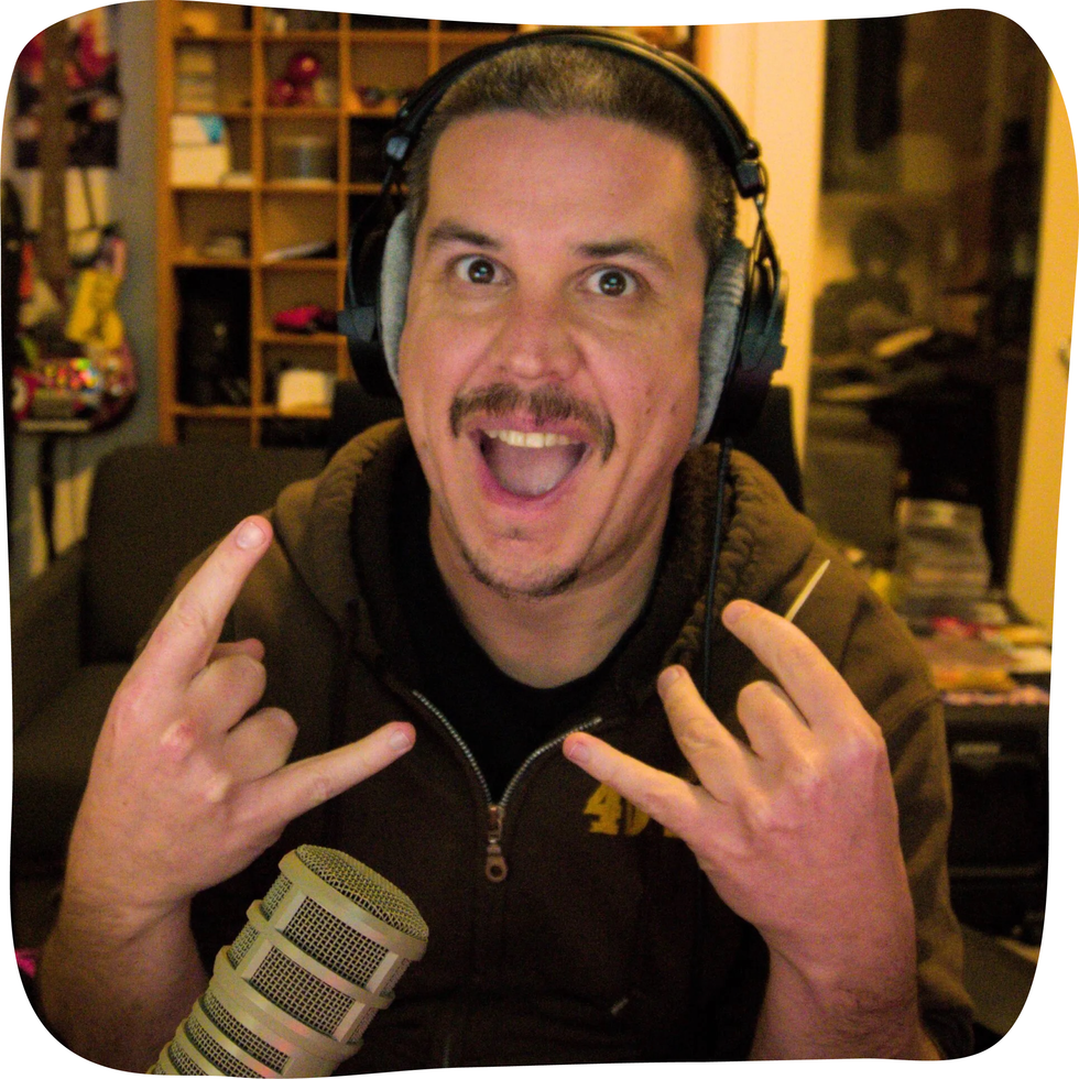 Man with headphones making rock-on hand gestures, smiling widely. Brown hoodie, microphone, and cluttered background. This is Nick