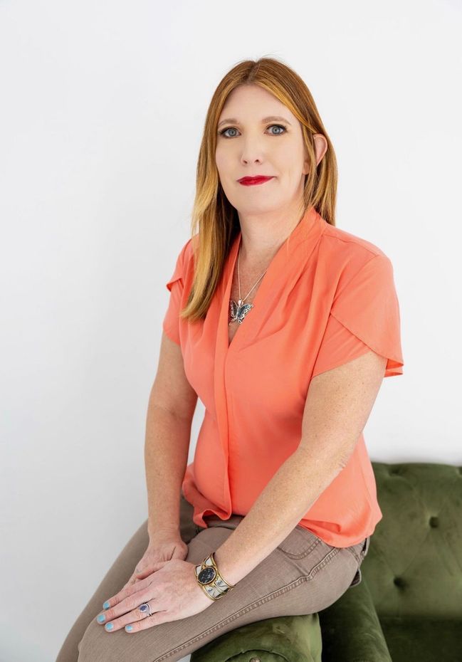 A person with long reddish hair wearing an orange top sits on a dark green velvet couch against a white background.