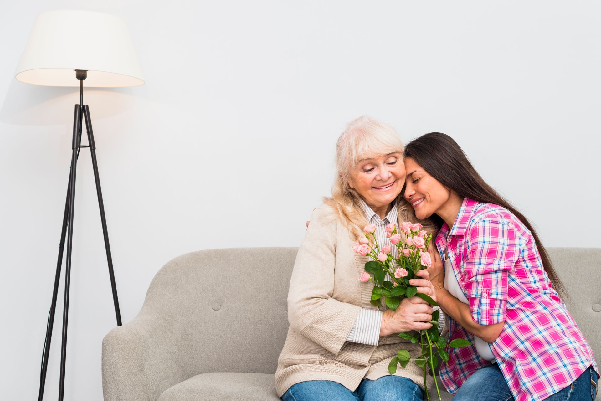 Woman in pink shirt hugs an older woman on a sofa, holding flowers beside a floor lamp.