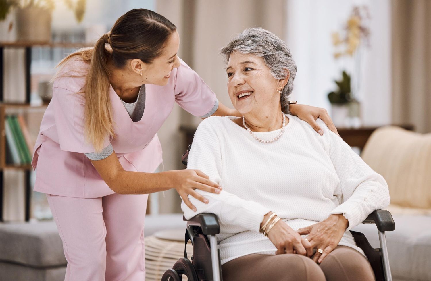 Nurse in pink scrubs comforts an elderly woman in a wheelchair, indoors.