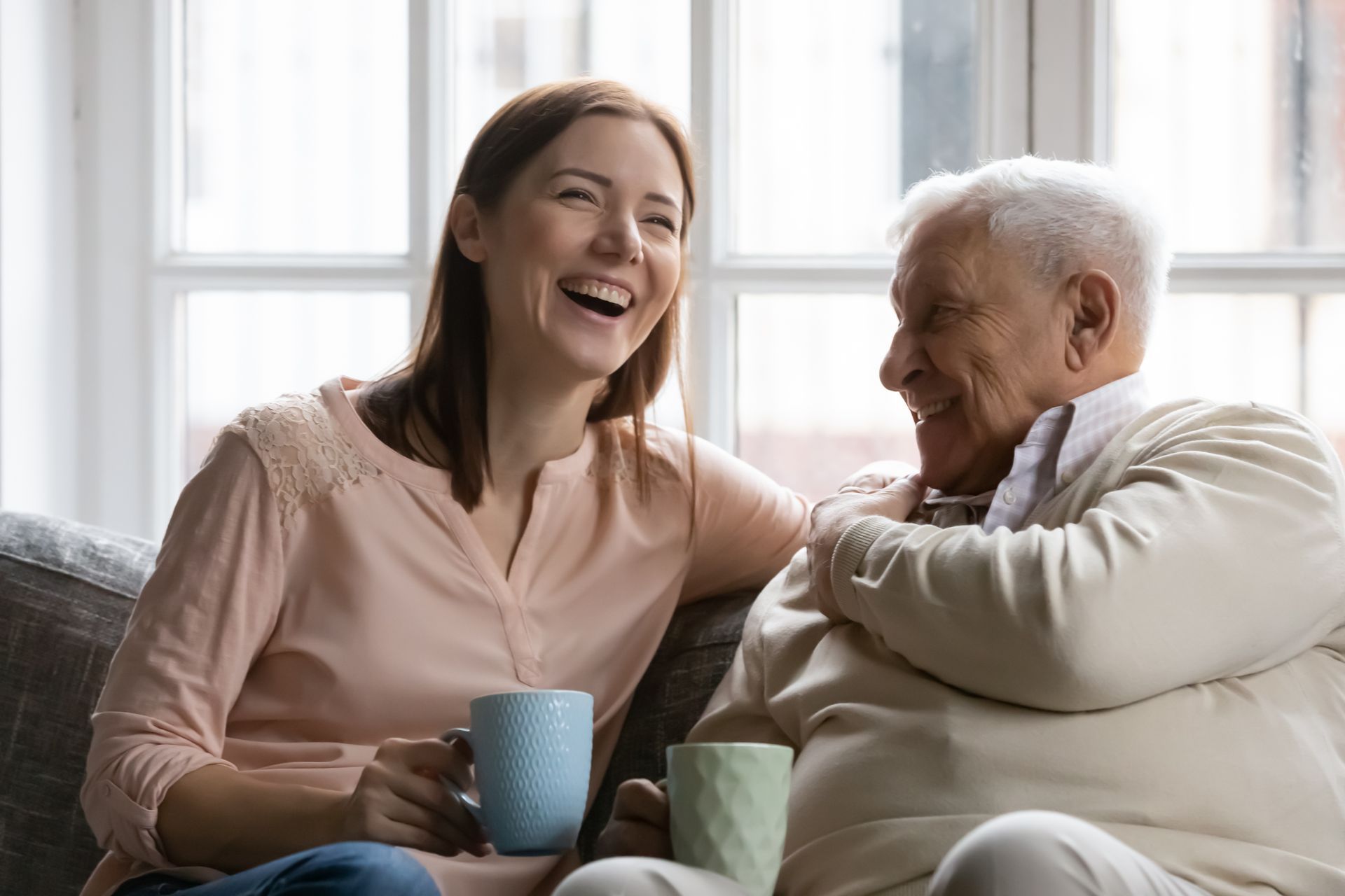 Woman and older man laughing, sitting together on a couch, holding mugs.