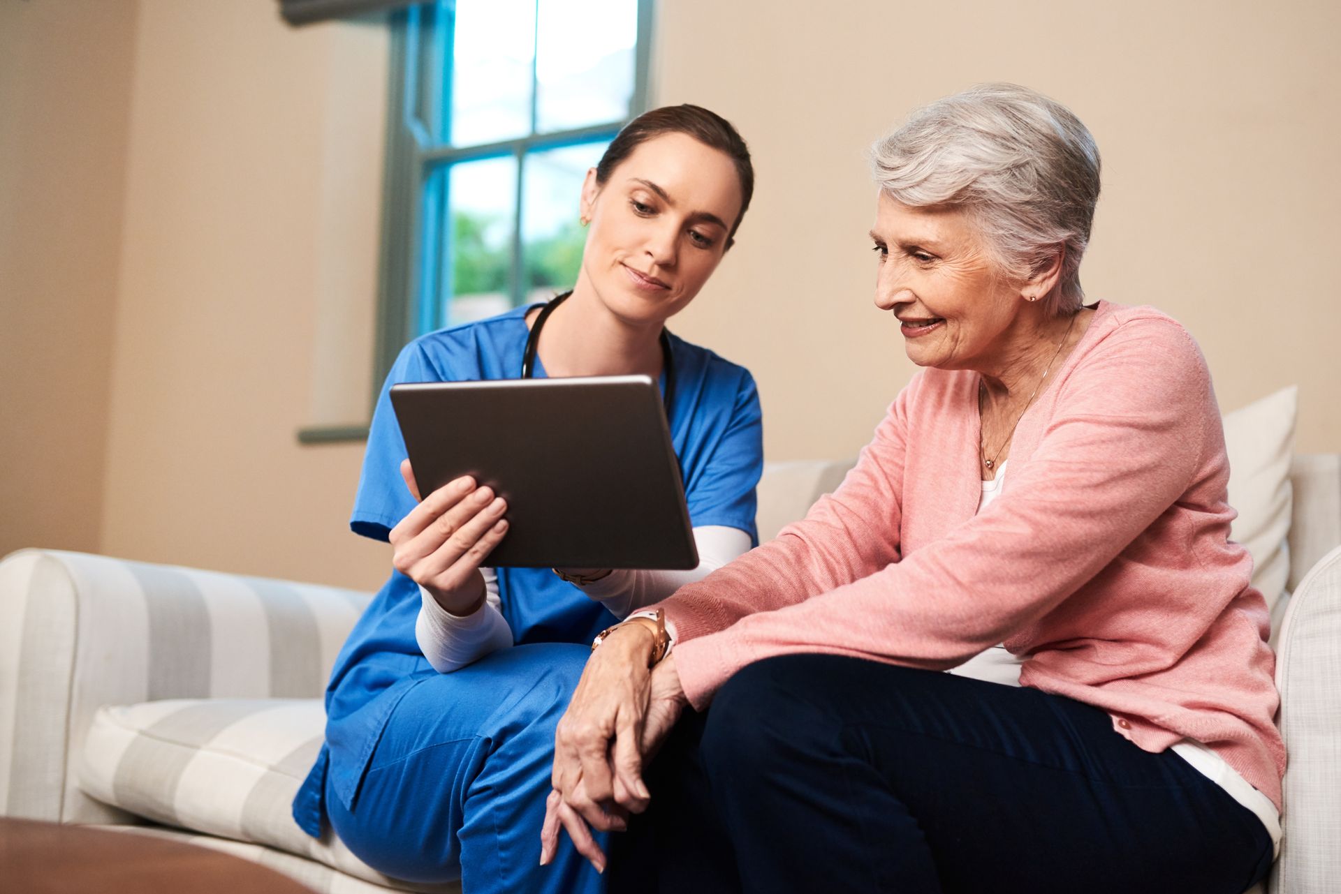 Nurse in blue scrubs shows tablet to elderly woman, both seated on sofa.