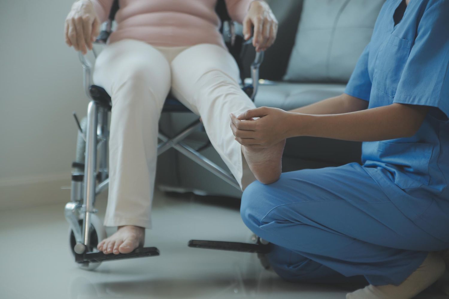 Nurse examining the foot of a person in a wheelchair; indoor setting, blue scrubs.