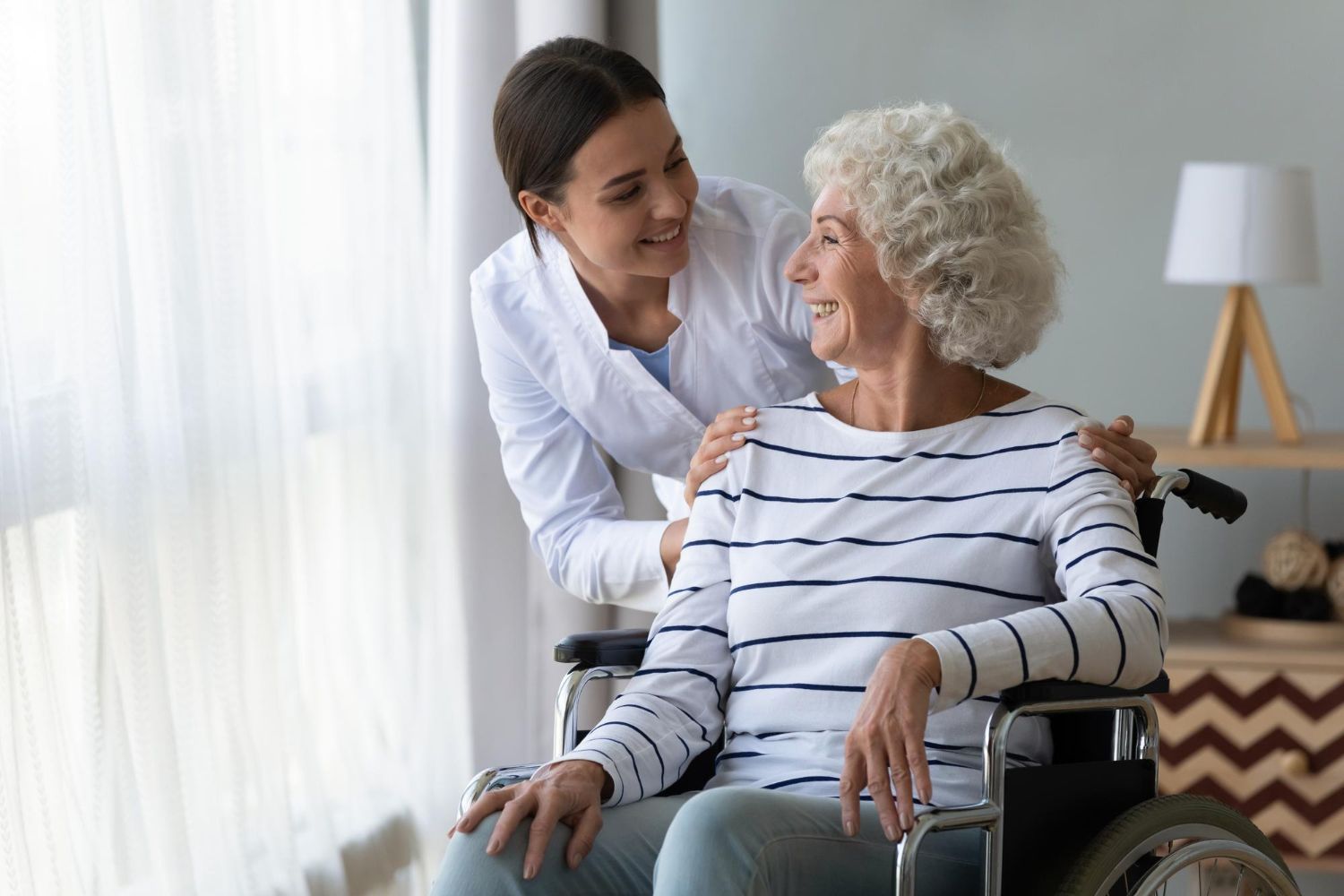 Woman in a wheelchair smiles at a caregiver with a hand on her shoulder, indoors.
