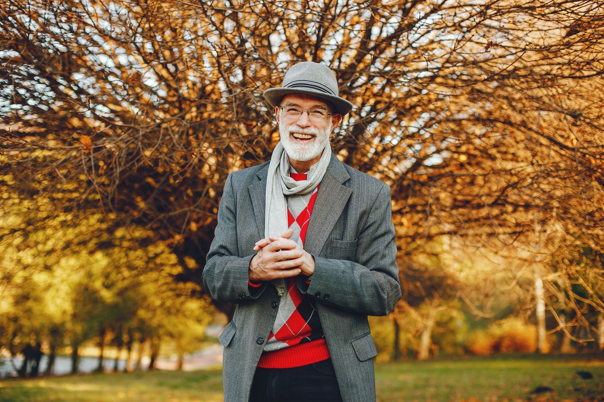 Man holding a coffee cup in a gray coat and hat, standing outdoors among autumn trees with warm sunlight