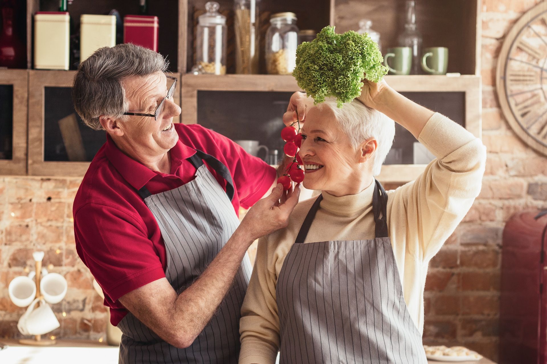 Elderly couple in kitchen playfully holding vegetables, smiling.
