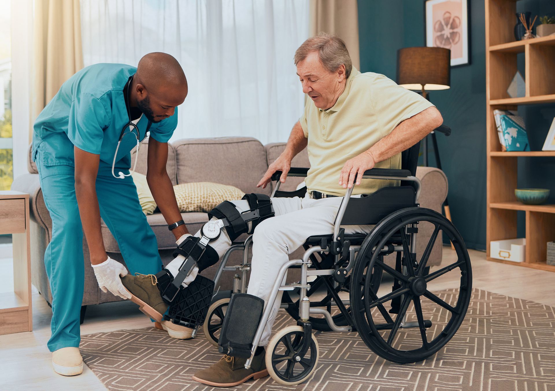 Caregiver assisting a person with a leg brace in transferring from a wheelchair in a living room.