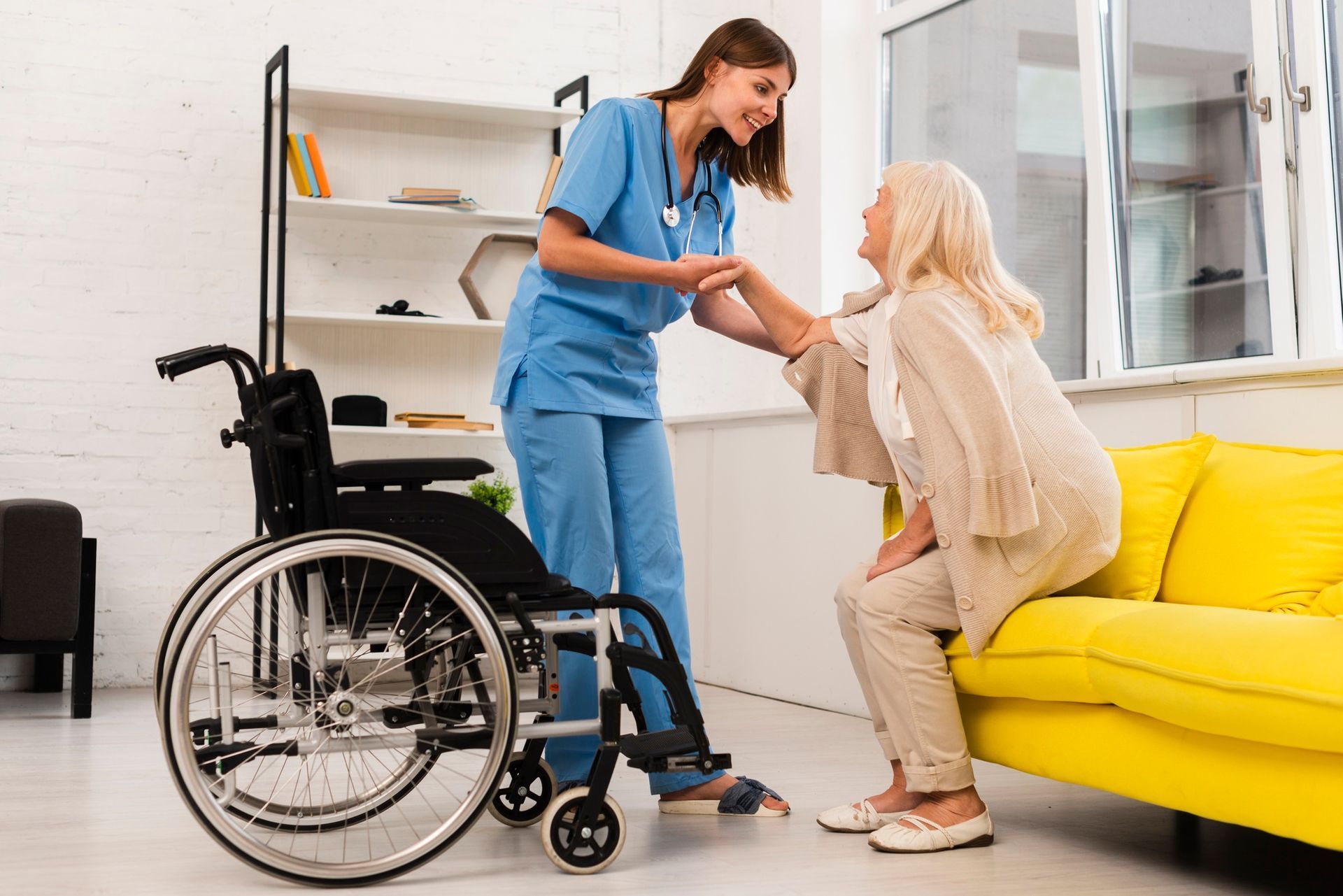 Nurse assists senior from a yellow couch to a wheelchair; indoor setting.
