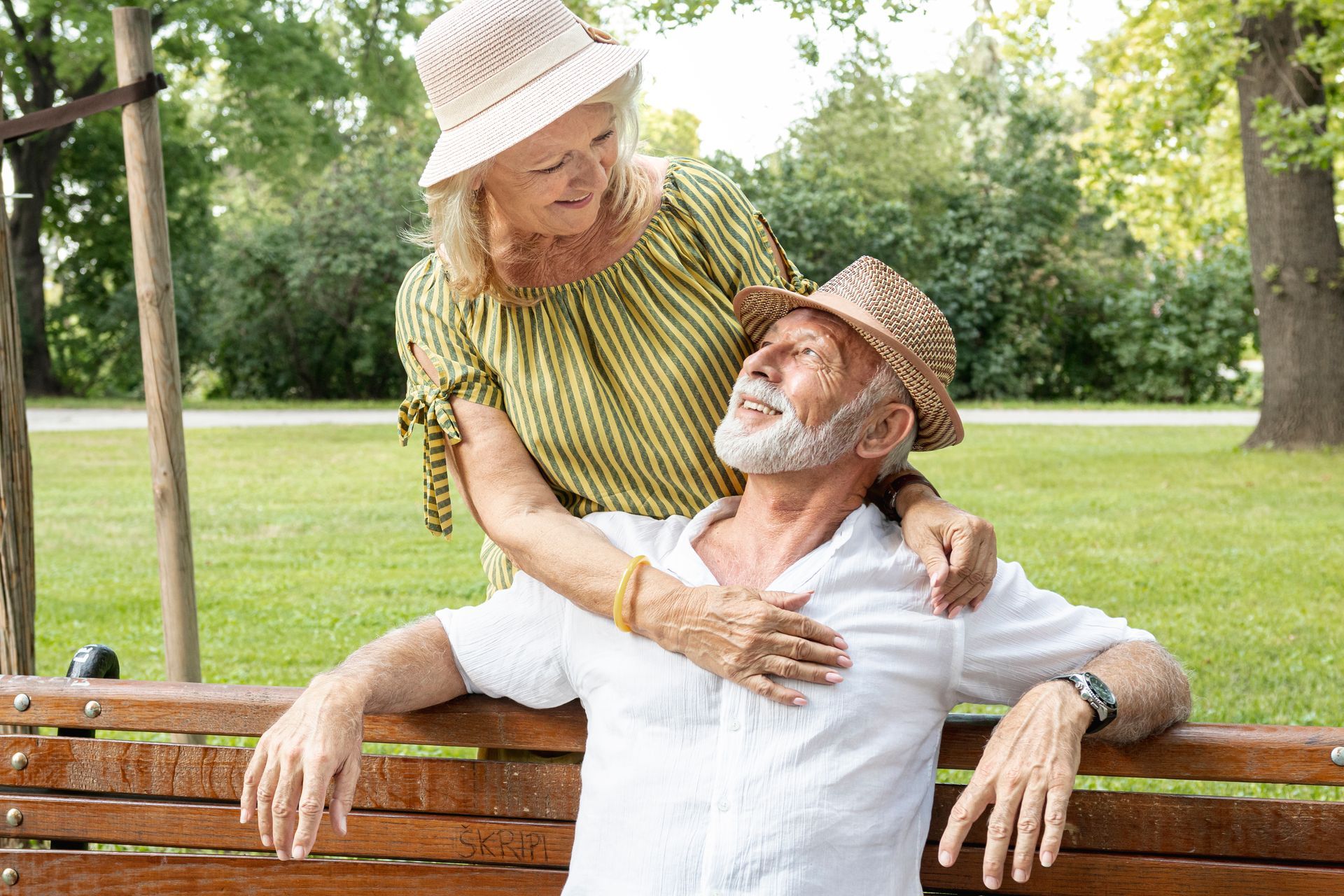 Two older adults smiling on a park bench, embracing in a sunny green park.