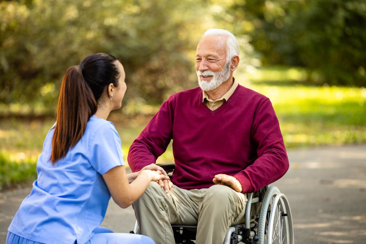 A person in a wheelchair smiles at a caregiver in a park. The caregiver holds their hands.