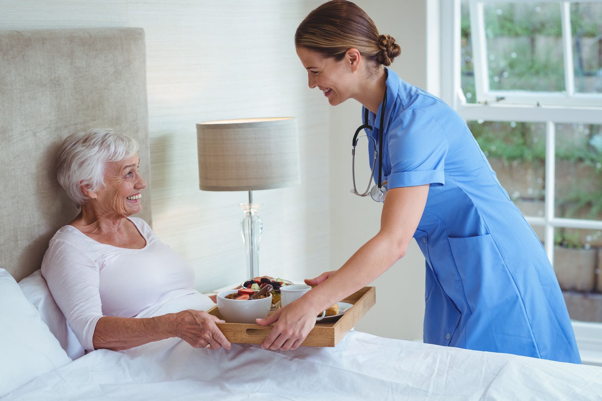 Nurse serving breakfast to a patient in bed; both smiling. Room with window and lamp.