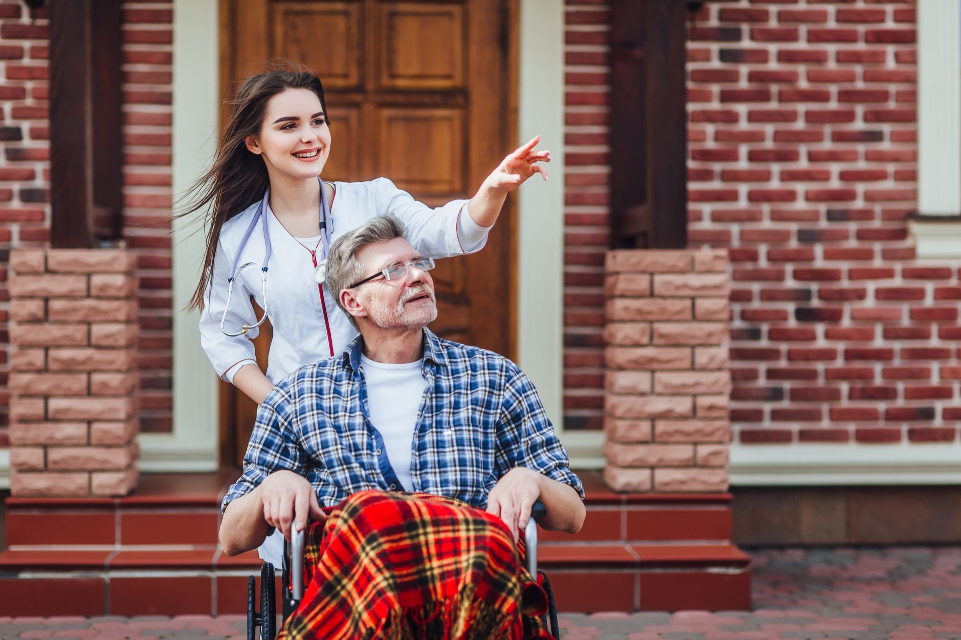 Young woman points ahead while helping an older person in a wheelchair outside a brick building