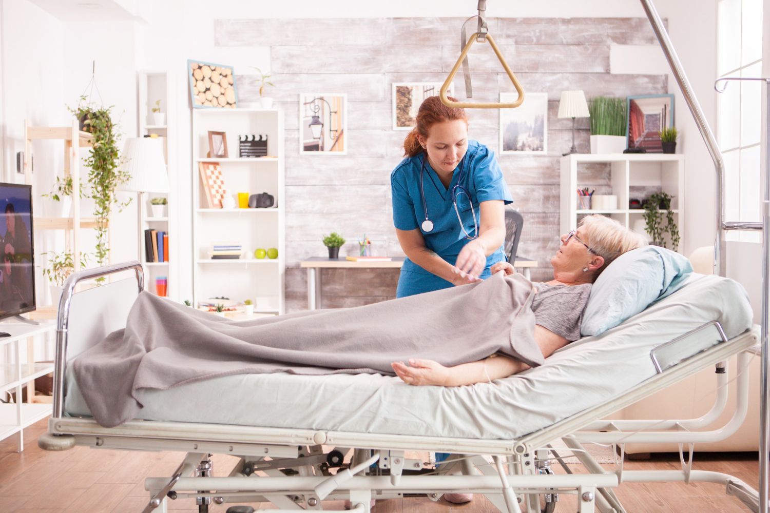 Nurse tending to an elderly patient in a home setting. Nurse wears blue scrubs. Patient lies in bed covered by a blanket.