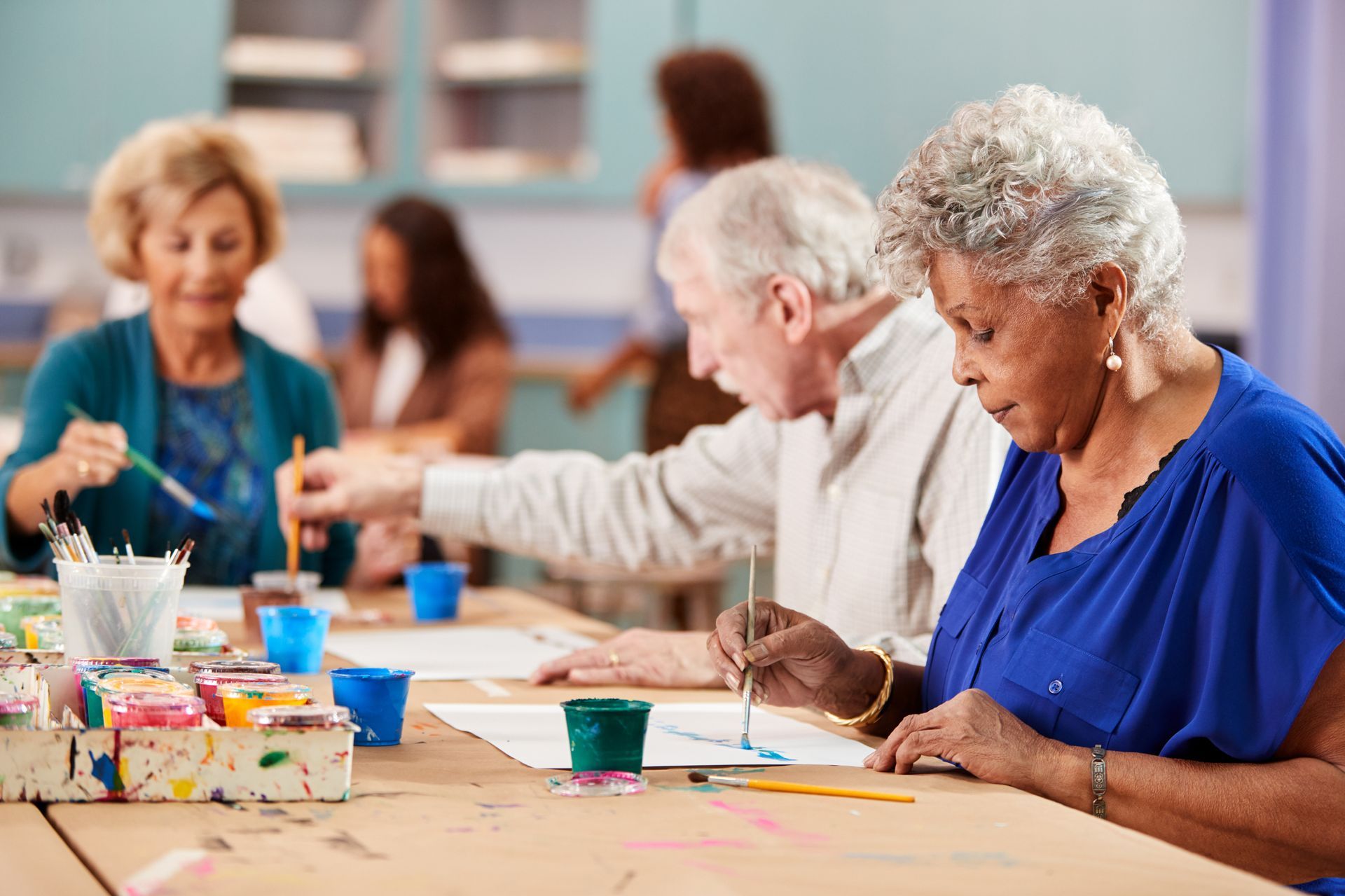Seniors painting in art class; diverse group at a table, using brushes and paints.