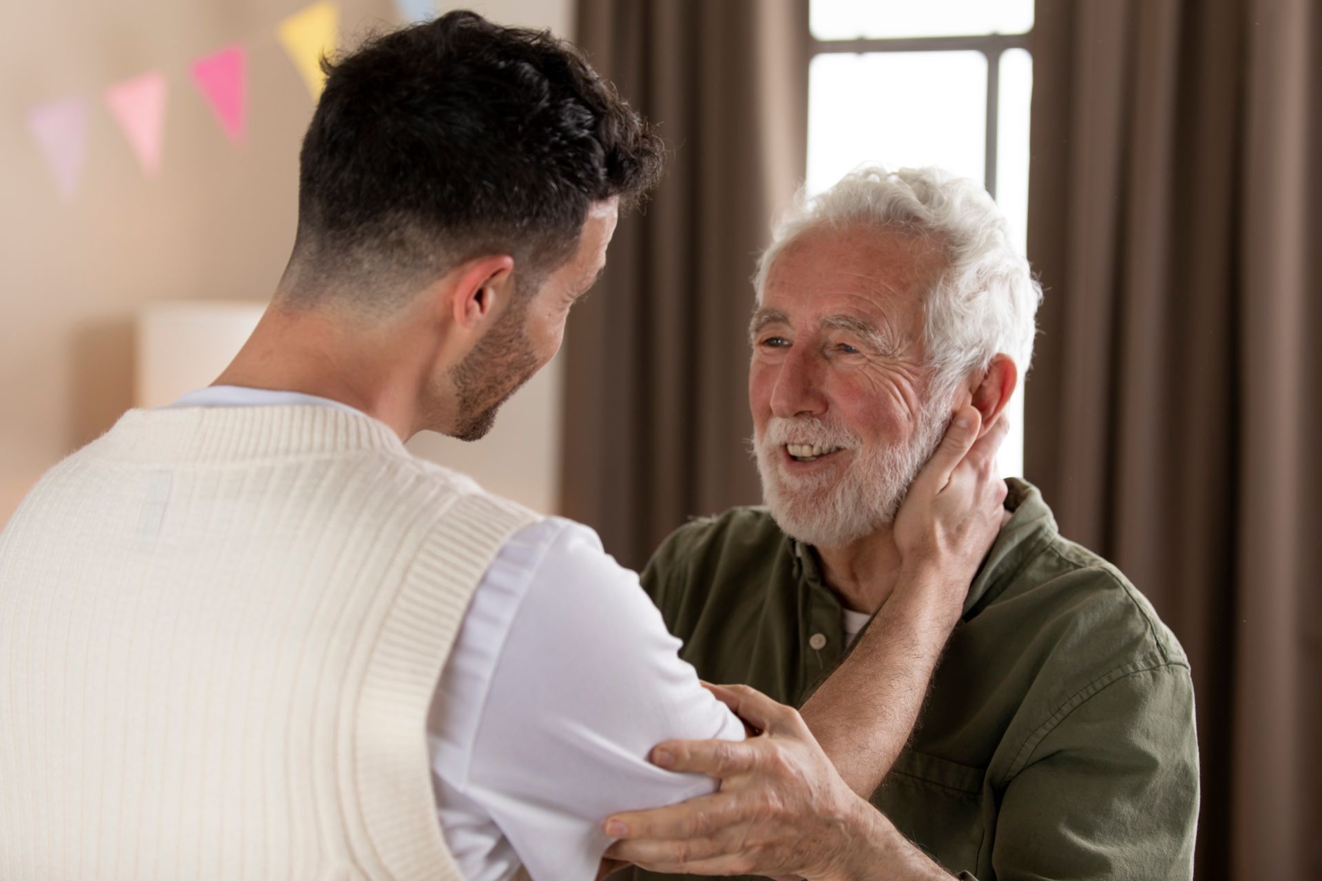 Two people embracing warmly in a bright indoor setting, one smiling with hand on the other's shoulder.