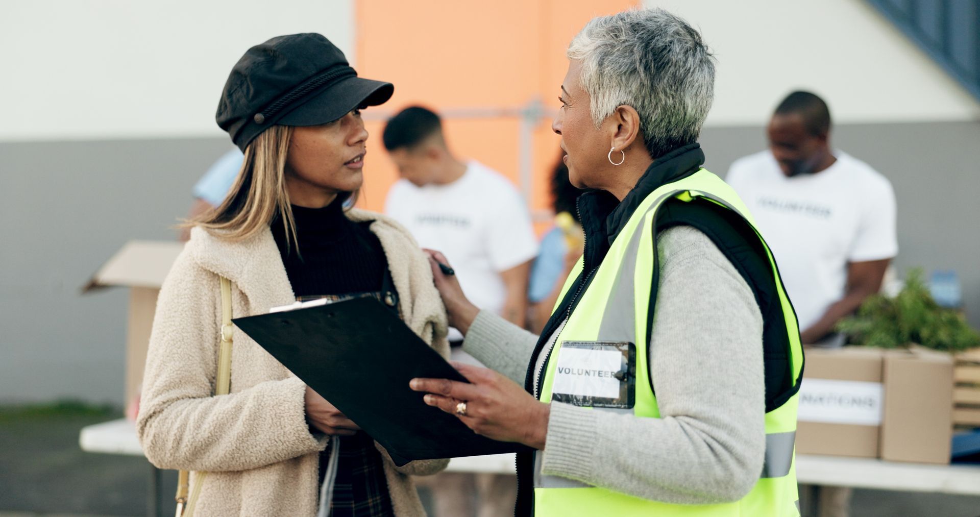Two women talking with a clipboard, one wearing a volunteer vest. Other volunteers and boxes in the background.