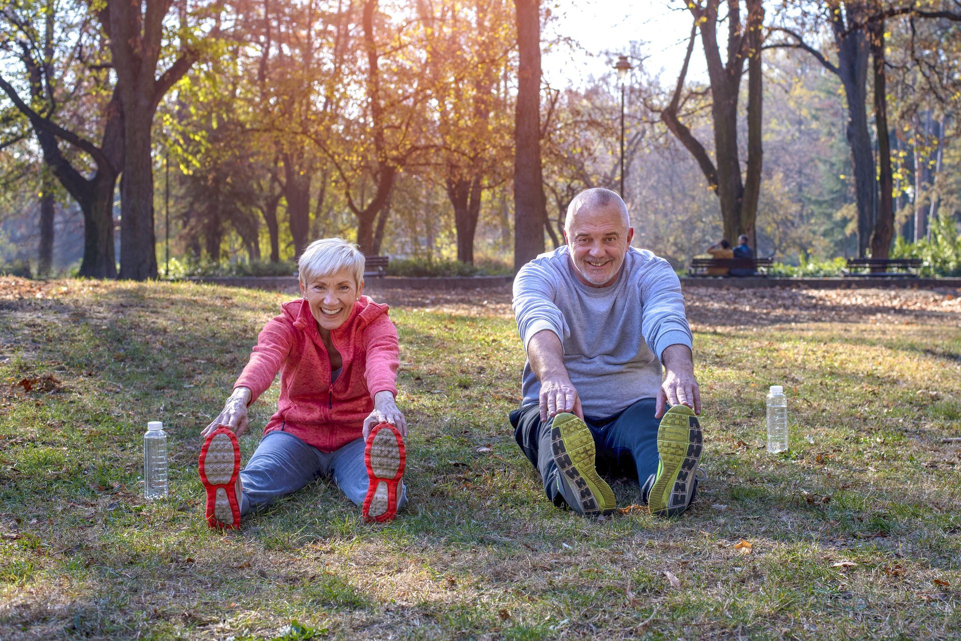 Two people stretch on grass in a park with trees and autumn light.