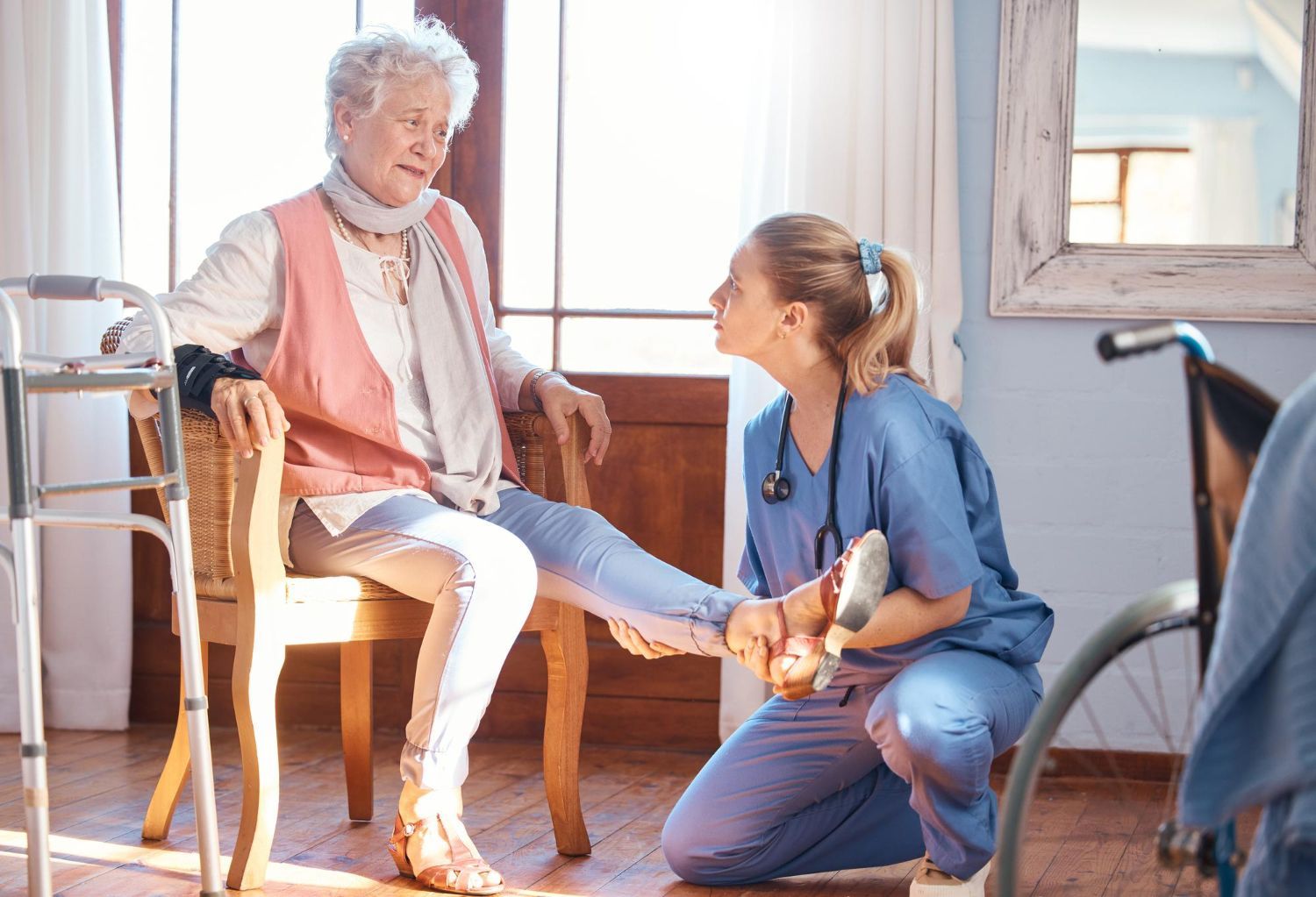 Nurse examining an older person's foot, indoors. Walker and wheelchair are present.