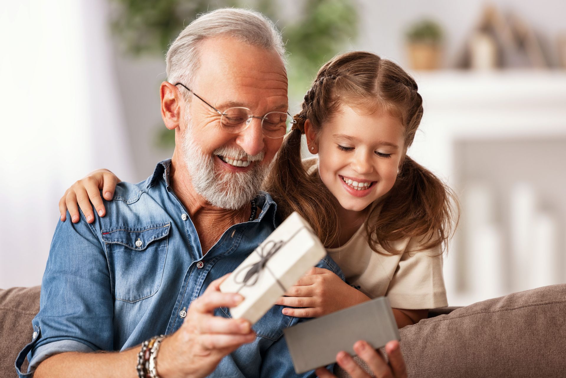 Grandfather and granddaughter open a gift box, smiling on a couch.