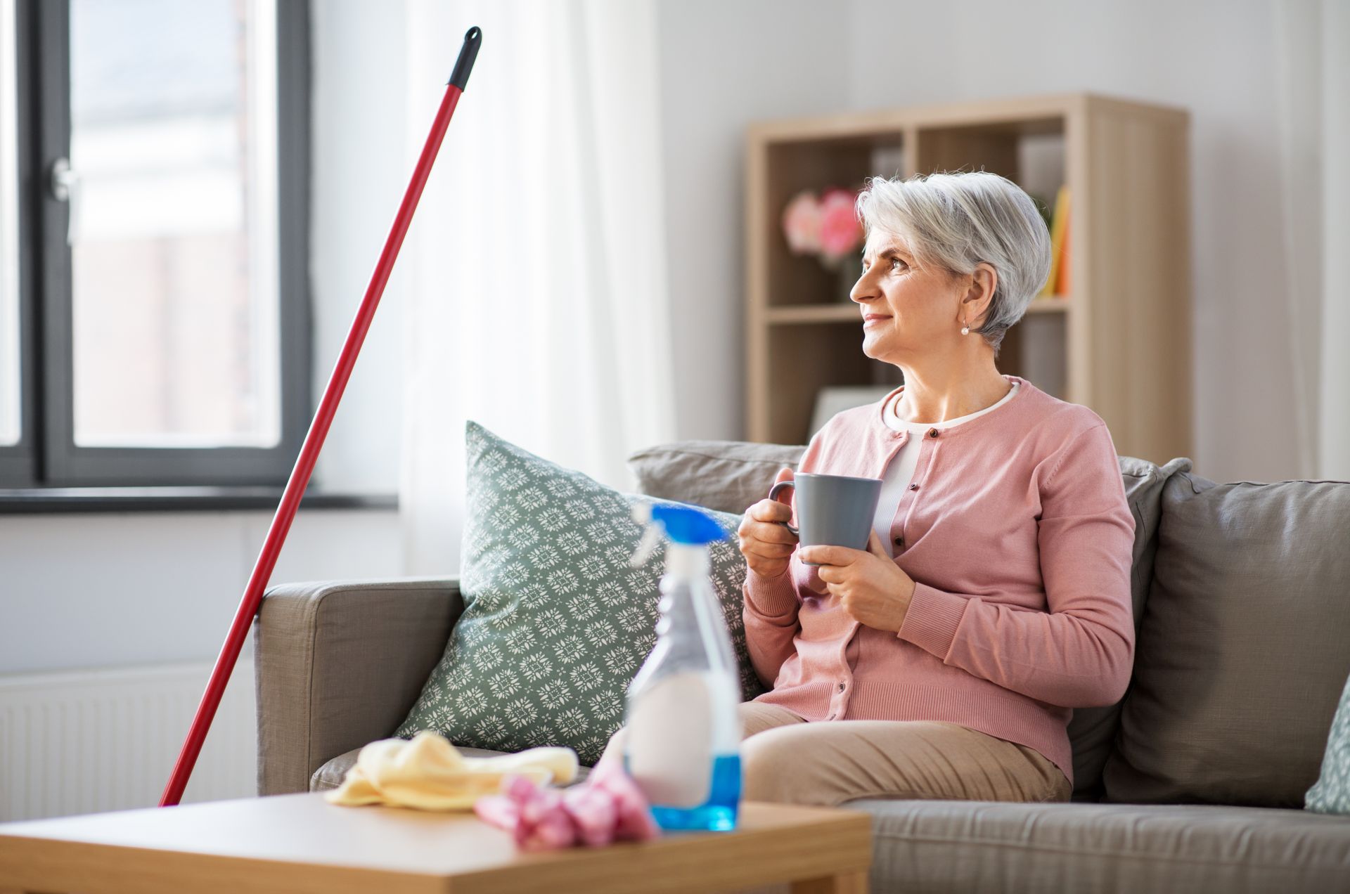 Woman sitting on a sofa in a bright living room, holding a cup beside cleaning supplies and a mop
