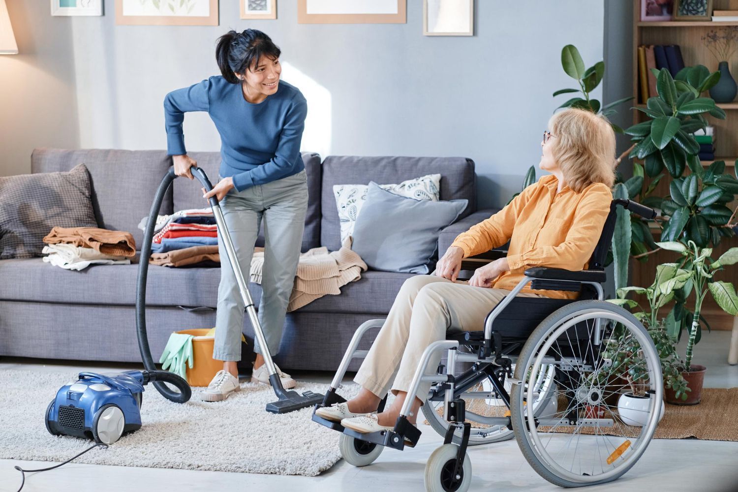 Woman vacuuming while talking to a person in a wheelchair in a living room.