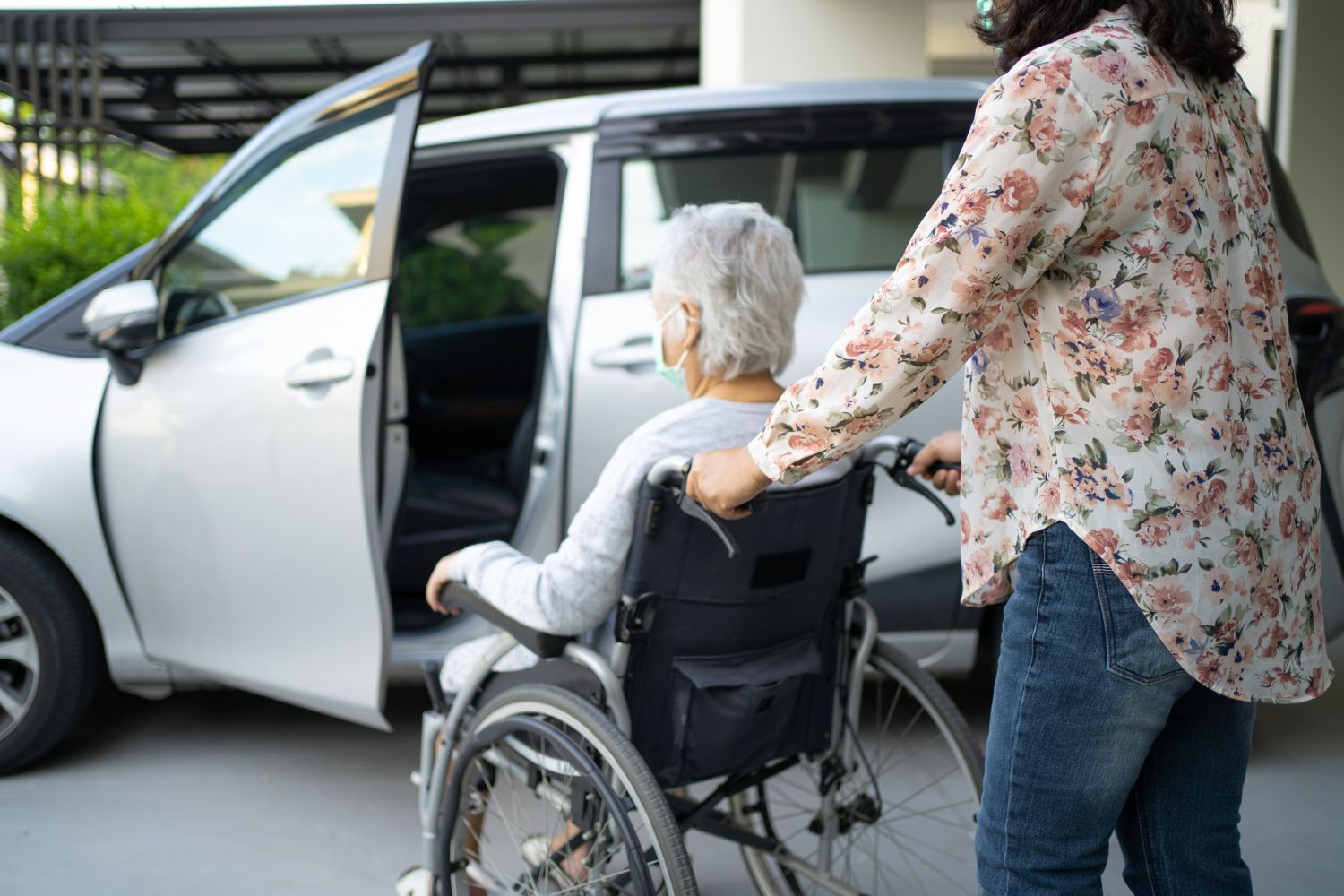 Woman assisting a person in a wheelchair toward a car with an open door.