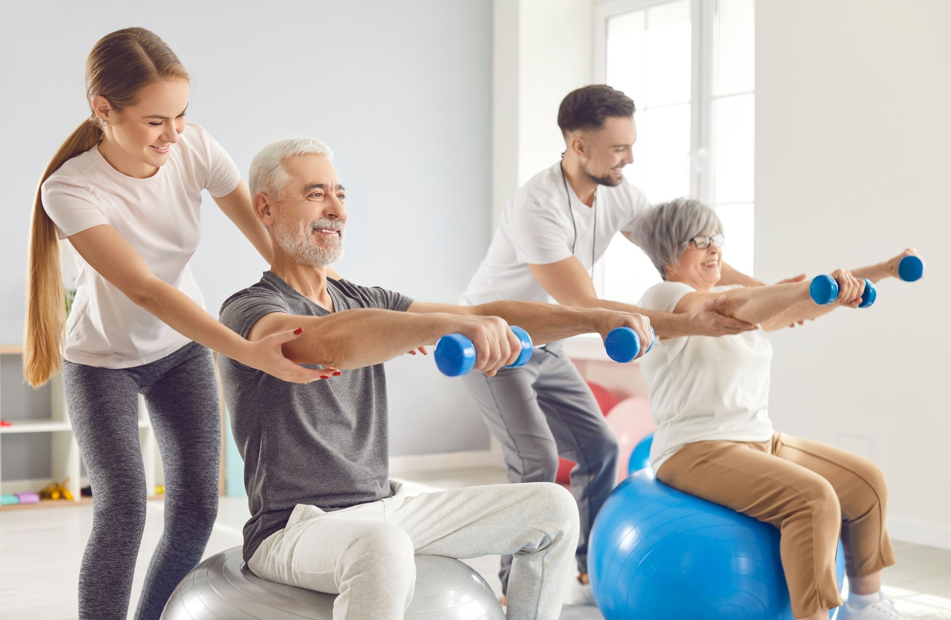 Two elderly people exercising with dumbbells on exercise balls, aided by two trainers. Indoors.