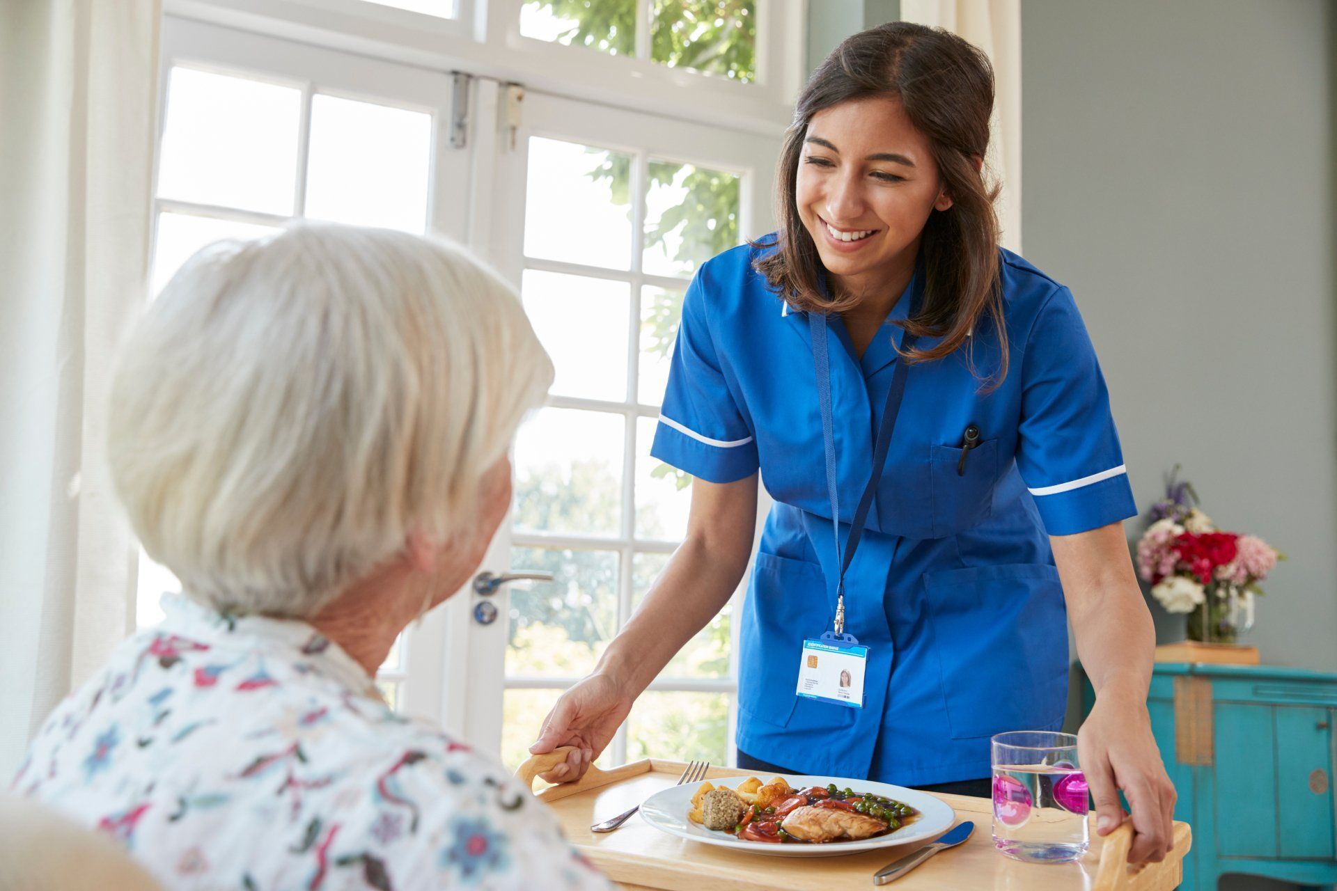 Caregiver serving a meal to a patient at a window-lit table. Both smiling; caregiver in blue uniform.