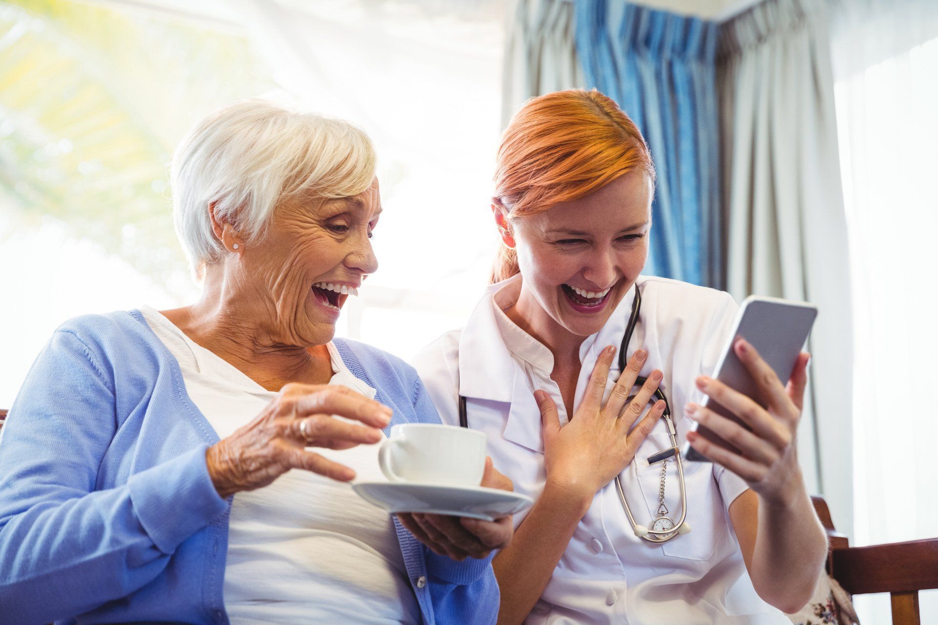 Elderly woman and caregiver laughing while looking at a smartphone, indoors with cup of tea.