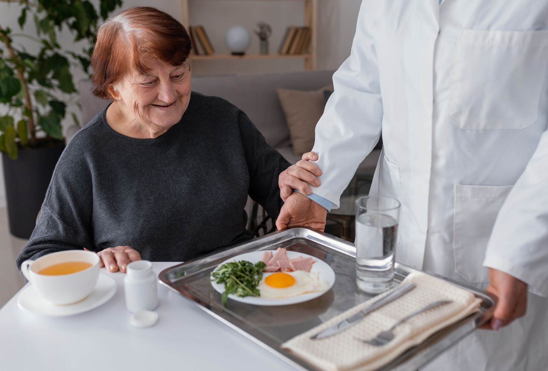 Woman smiles as a medical professional offers a tray with food and water. Indoor setting, white table.