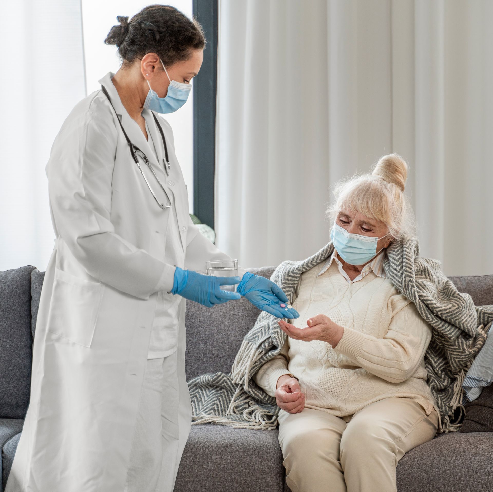 Healthcare worker handing medication to a person wrapped in a blanket, both wearing masks.