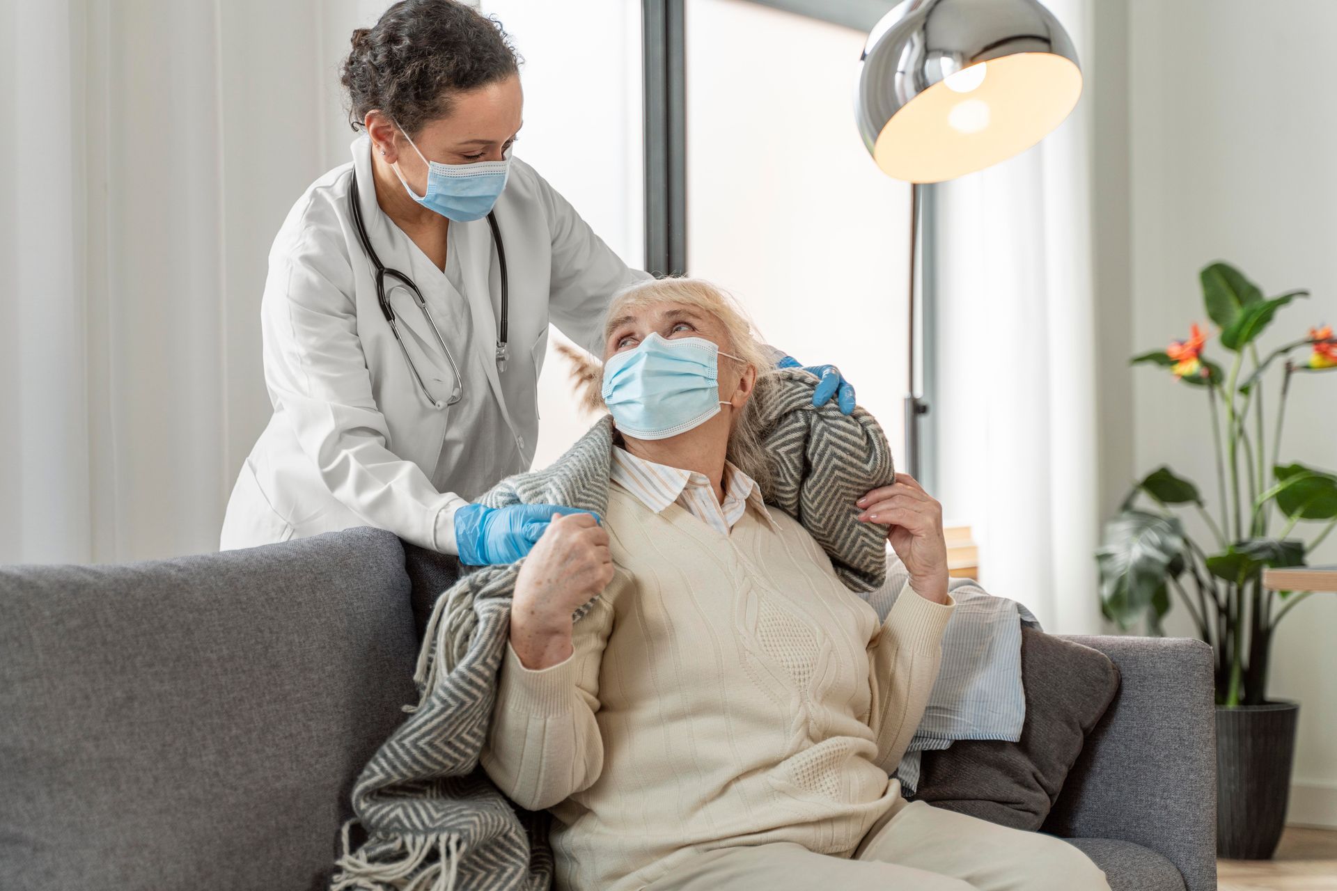 Healthcare worker assists elderly person, both wearing masks, with a blanket indoors.