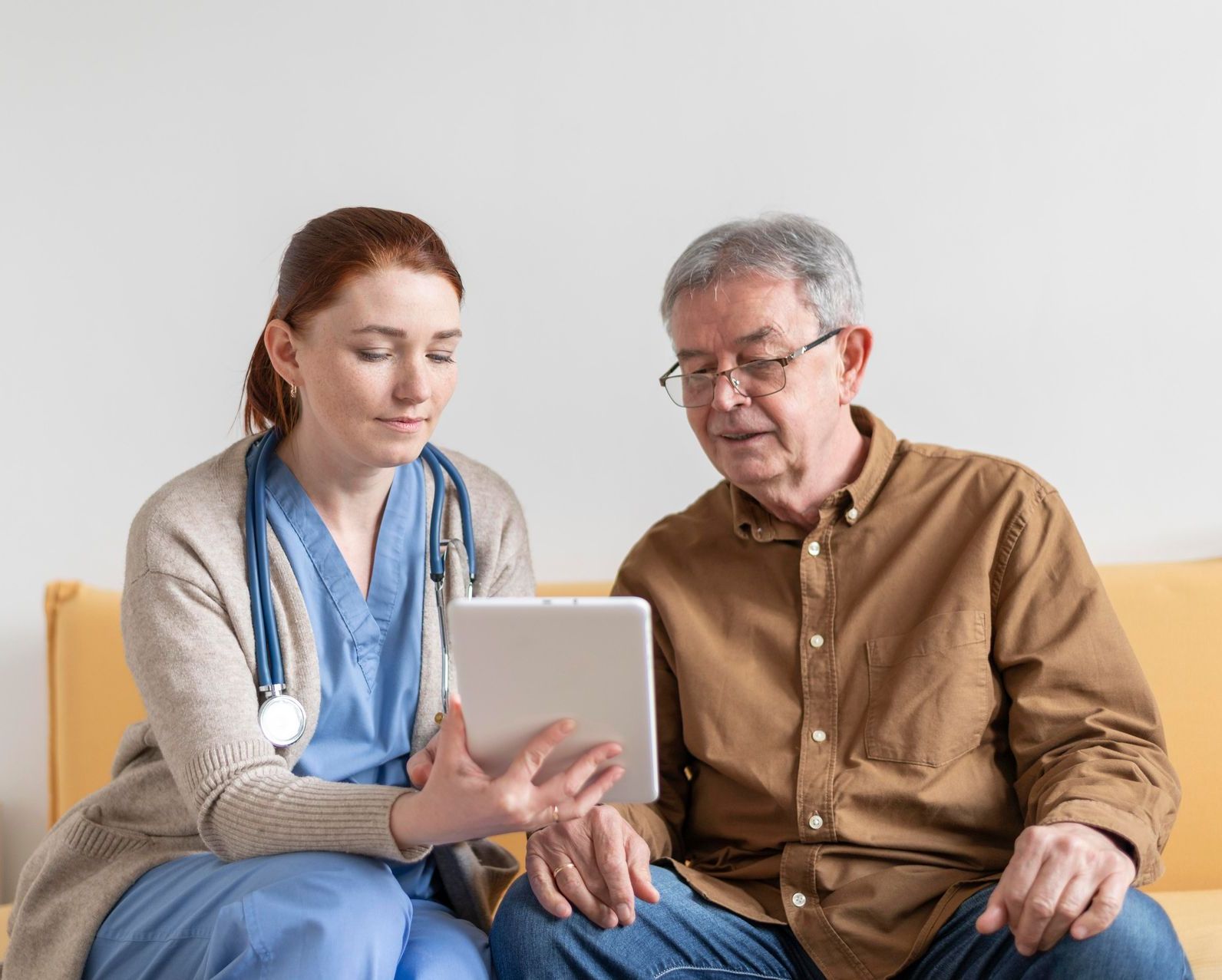 A doctor and an elderly man are sitting on a couch looking at a tablet.