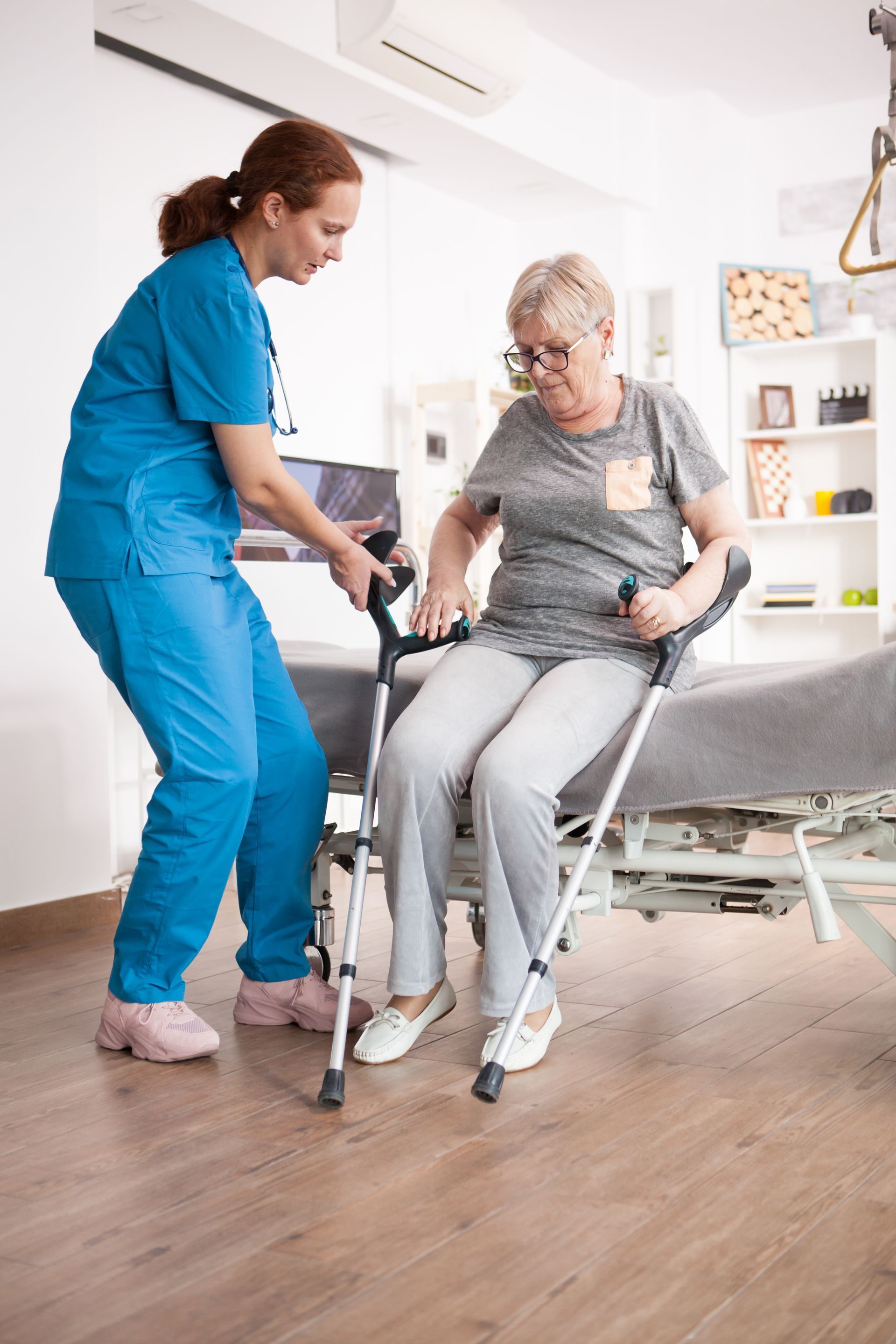 Nurse helping a patient with crutches to sit. Indoor setting with medical equipment.